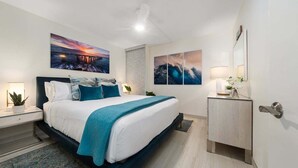 Moderni hotellihuone, jossa on suuri sänky, yöpöytä lampun kanssa, sivupöytä, jossa on kasvi, ja taideteoksia seinillä.