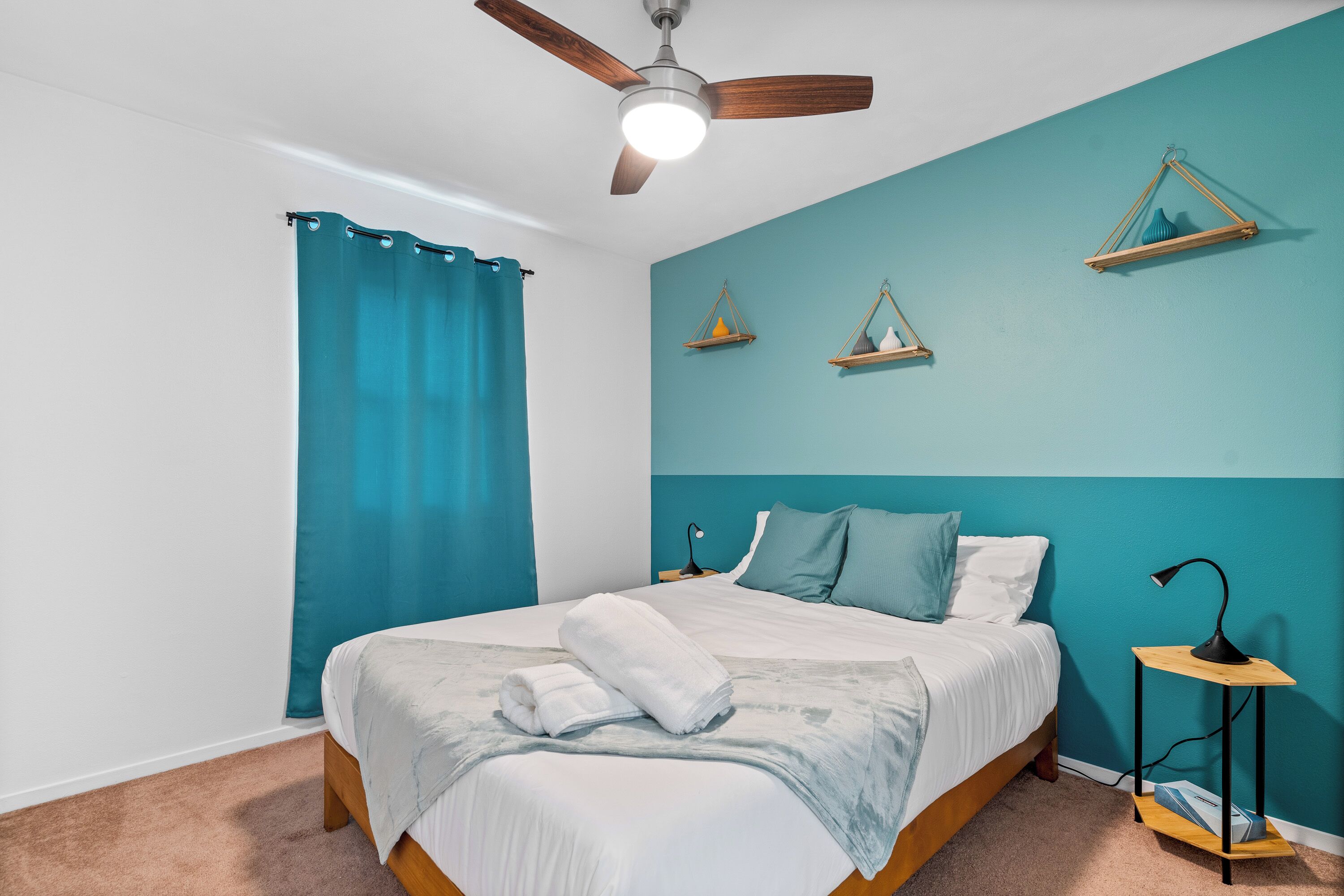 Un dormitorio con cama, ventilador de techo, mesita de noche y ventana con cortinas.