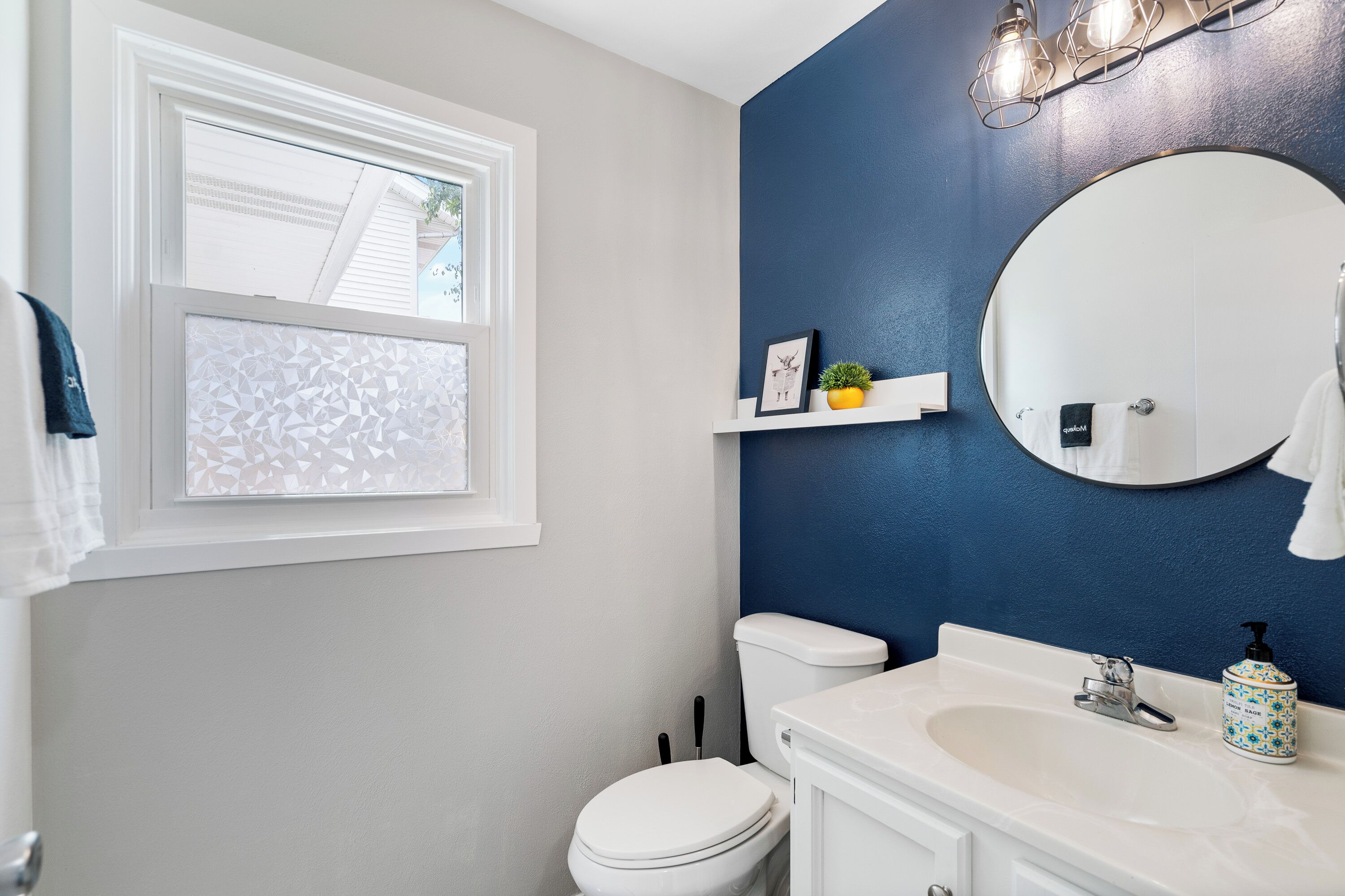 A bathroom with a white toilet, a sink with a soap dispenser, a mirror, and a window with frosted glass.