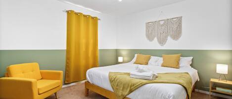A bedroom with a bed, a yellow armchair, a yellow curtain, a macrame wall hanging, and a bedside table with a lamp.