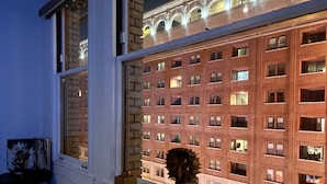 A room with a large window overlooking a brick building with multiple windows. There is a wooden table with a sculpture and a magazine on it. - Iconic Heart of DTLA Loft with Rooftop Pool/Jacuzzi/Skyline (Los Angeles)