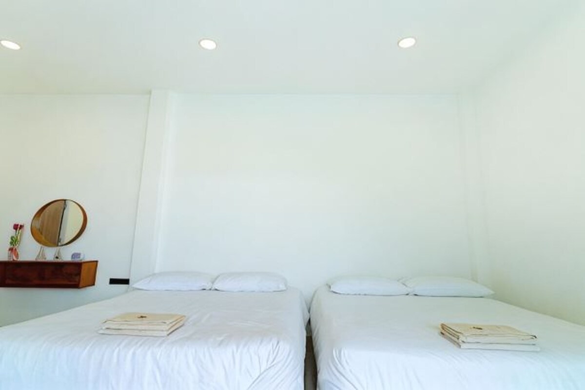 Una habitación con dos camas, un pequeño estante de madera con un espejo y flores, y una pared blanca lisa.