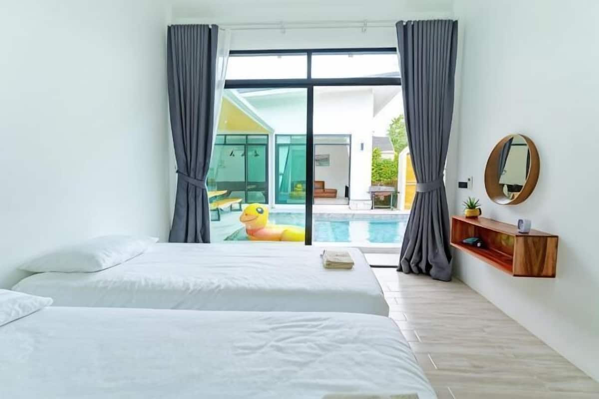 Quarto de hotel com duas camas, uma grande porta de vidro deslizante para uma área de piscina, uma prateleira de madeira com espelho e uma pequena planta.
