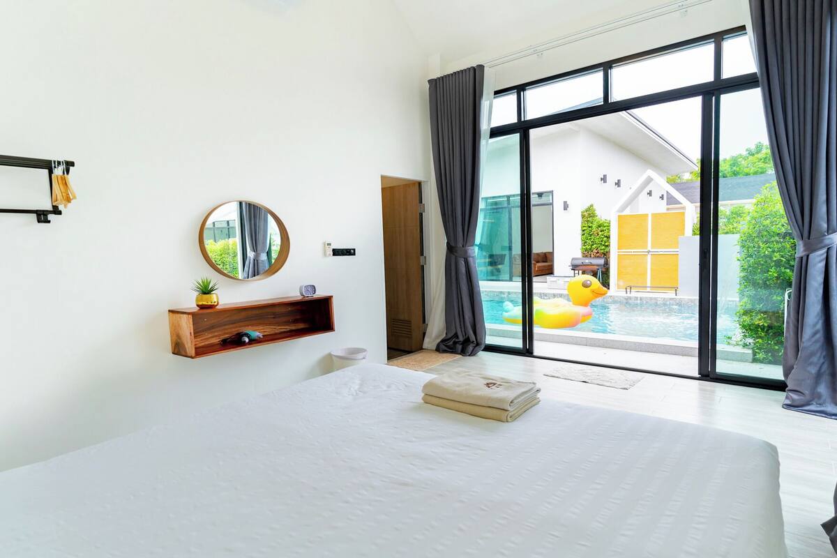 Quarto moderno com cama grande, prateleira de madeira com planta, espelho redondo e vista para piscina e área externa.