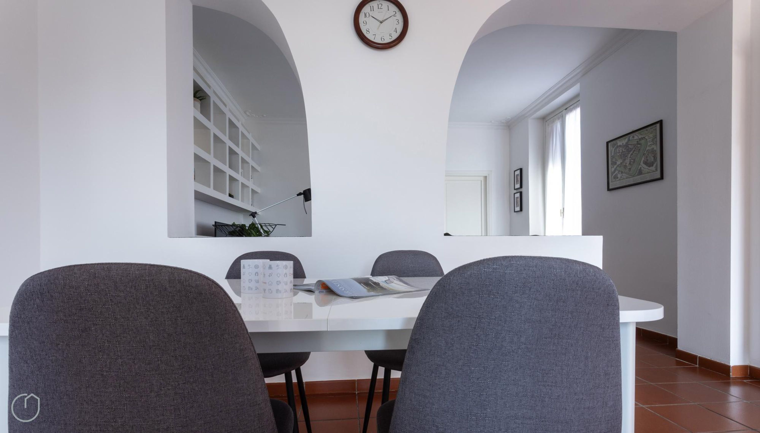 Un'area pranzo con un tavolo bianco, due sedie grigie, una sveglia appesa al muro e un quadro incorniciato.