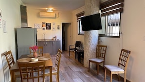 A dining area with a wooden table and chairs, a kitchen in the background, and a flat-screen TV mounted on a pillar. - Chalet in Batrouna Park resort, facing the pool, gazon,gym,GENERATOR IS INCLUDED (Helweh)