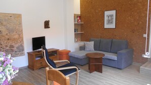 A living room with a stone wall, a grey sofa, a wooden TV stand, a wooden chair, and a small round table. - Cottage with 2 bedrooms with both en-suite bathrooms in a seaside village (Salir do Porto)