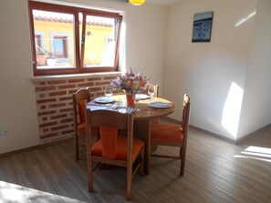 Uma área de refeições com parede de tijolos, mesa de madeira, cadeiras laranja e uma janela com vista para um edifício.