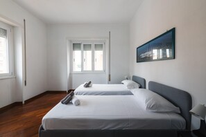 Quarto de hotel com duas camas de solteiro, uma janela grande com persianas, um quadro na parede e piso de madeira.