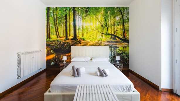 A bedroom with a large forest mural on the wall, a bed with white linens, and a wooden floor.