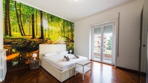 A bedroom with a large forest mural, a white bed, a small table, and a sliding glass door to a balcony.