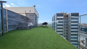 A rooftop terrace with artificial grass, a seating area, and a glass railing overlooking a cityscape.