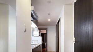 A narrow hallway with wooden flooring, a kitchen area with white appliances, and a dark door. - Modern 1Br Apt near Hondori District for 6ppl (Naka Ward, Hiroshima)