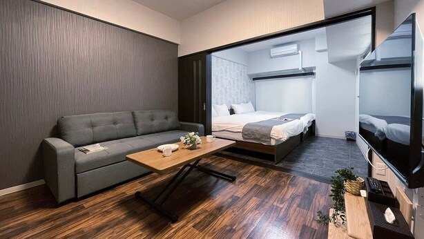 A compact hotel room with a sofa, a bed, a TV, and a wooden coffee table. - Modern 1Br Apt near Hondori District for 6ppl (Naka Ward, Hiroshima)