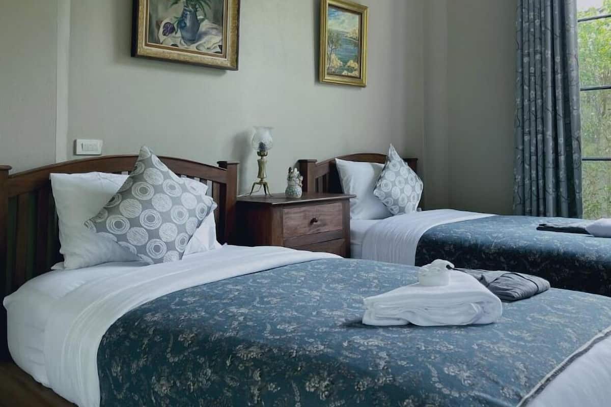 A hotel room with two beds, each with a blue floral bedspread, white pillows, and a nightstand with a lamp. There are two framed paintings on the wall, and a window with curtains.