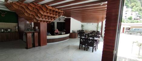 A spacious lobby with a wooden ceiling and a large, abstract sculpture. There is a reception desk and a seating area with tables and chairs.