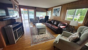 A spacious living room with a large flat-screen TV, a leather sofa, a coffee table, and a view of a lake through large windows. - Lakefront House with dock and lots of toys! (Stanwood)