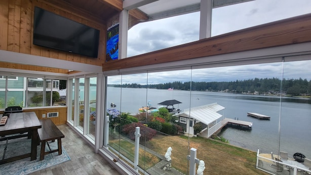 A large room with a TV, a dining table, and a view of a lake with boats and a dock. - Lakefront House with dock and lots of toys! (Stanwood)
