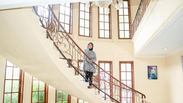 A person on a curved staircase with ornate metal railings, large windows, and a ceiling with hanging light fixtures. - Jalan Bagindo Aziz Chan (Saringan)