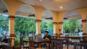A covered outdoor seating area with tables and chairs, surrounded by columns and greenery. - Jalan Bagindo Aziz Chan (Saringan)