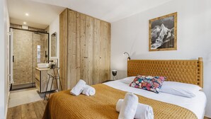 Een slaapkamer met een houten hoofdbord, een bed met witte lakens en een kleurrijk kussen, een badkamer met een douchecabine van glas, en een lijst met een foto aan de muur.