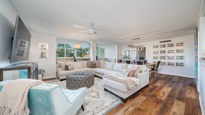 A modern living room with a sectional sofa, a tufted ottoman, a television, and a wall-mounted shelf with books. - Lakefront Retreat: Updated Condo with Resort Amenities, Lake Views, Pool & Close to Hot Springs Fun! (Hot Springs)