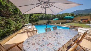 Ein Poolbereich mit einem Tisch, bedeckt mit einer gemusterten Tischdecke, umgeben von Holzstühlen und einem großen Sonnenschirm.