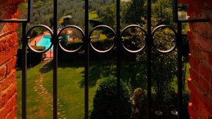 Vista a través de un arco de ladrillo con una reja metálica, que da a un paisaje verde con una piscina y edificios a lo lejos.