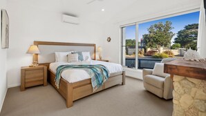 A bedroom with a bed, bedside tables, a nightstand, a chair, a stone accent wall, a ceiling fan, and a large window with a view of the outdoors. - Bella Beach Resort - An Oasis on the Bellarine (Saint Leonards)