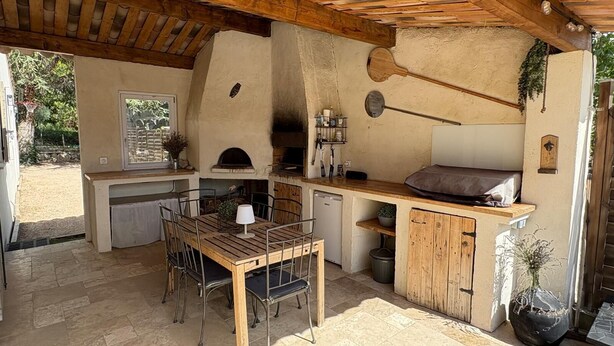 An outdoor kitchen with a stone countertop, wooden table, and chairs, a pizza oven, and a hanging light fixture.