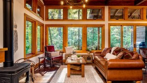 A cozy cabin interior with a fireplace, wooden furniture, and large windows offering a forest view. - Magnificent Mountain Cabin on 5 Acres Next to Creek, Picturesque Outdoor Space, Hot Tub, Ping Pong (Rhododendron)