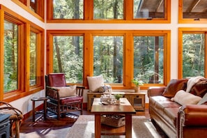 A cozy cabin interior with large windows, wooden furniture, and a view of a forest.