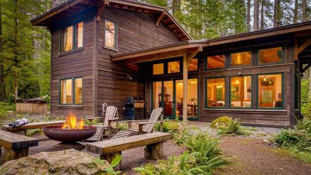 A rustic cabin with a fire pit, outdoor seating, and a large window overlooking a forested area. - Magnificent Mountain Cabin on 5 Acres Next to Creek, Picturesque Outdoor Space, Hot Tub, Ping Pong (Rhododendron)