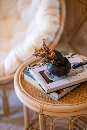 A wicker chair with a cushion, a small table with a woven surface, a blue ceramic pot with dried flowers, and a magazine. - Bi Ada Otelleri (Bozcaada)