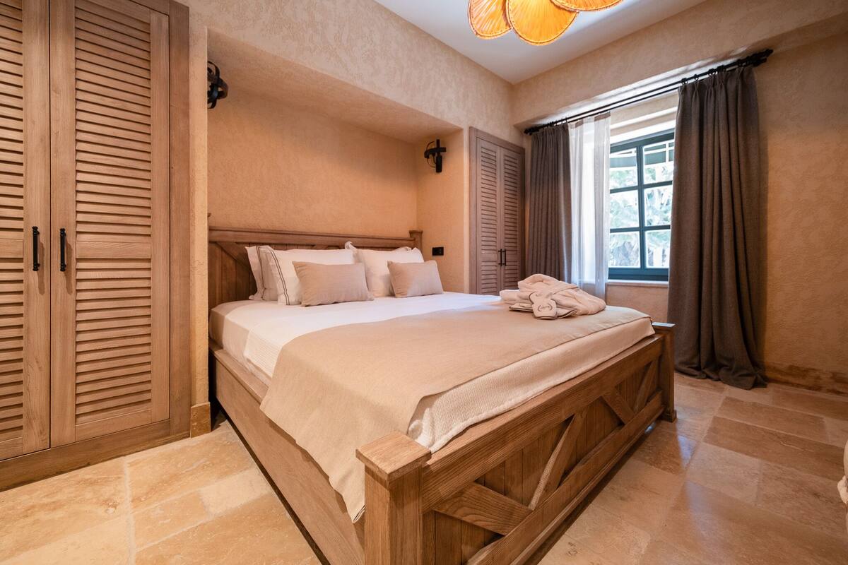 A bedroom with a wooden bed frame, beige bedding, a nightstand, and a window with curtains.