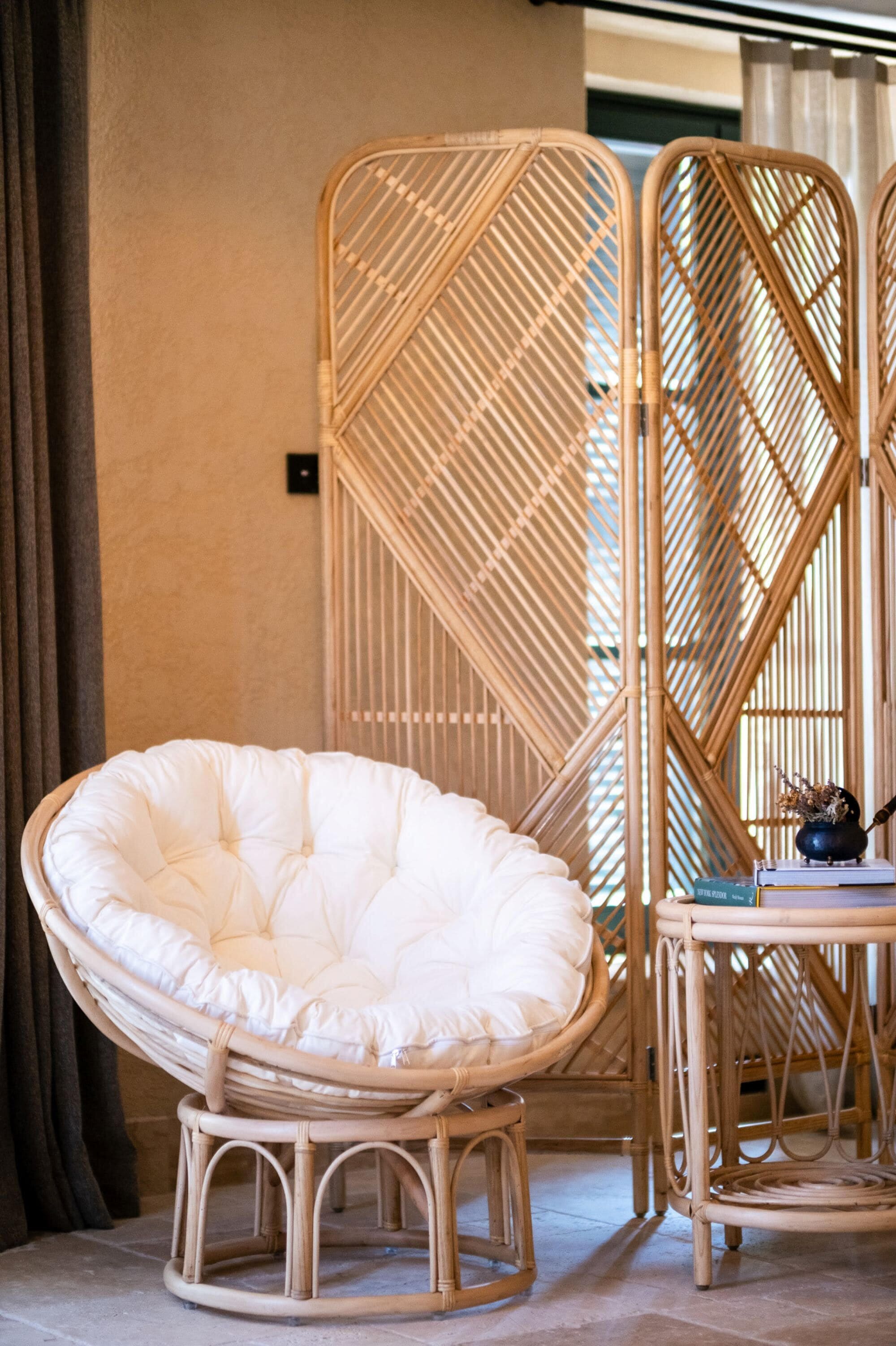 Una silla de mimbre con asiento y respaldo acolchados, una mesita con un libro y un objeto decorativo, y una partición de bambú en una habitación con suelo de baldosas y una cortina.