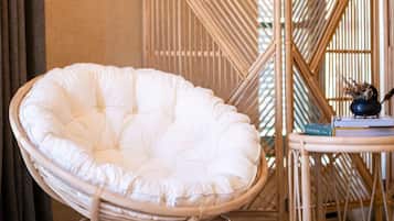 Una silla de mimbre con asiento y respaldo acolchados, una mesita con un libro y un objeto decorativo, y una partición de bambú en una habitación con suelo de baldosas y una cortina.