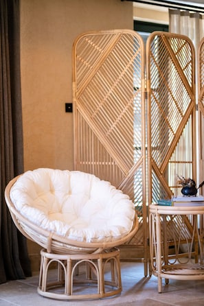 Una silla de mimbre con asiento y respaldo acolchados, una mesita con un libro y un objeto decorativo, y una partición de bambú en una habitación con suelo de baldosas y una cortina.