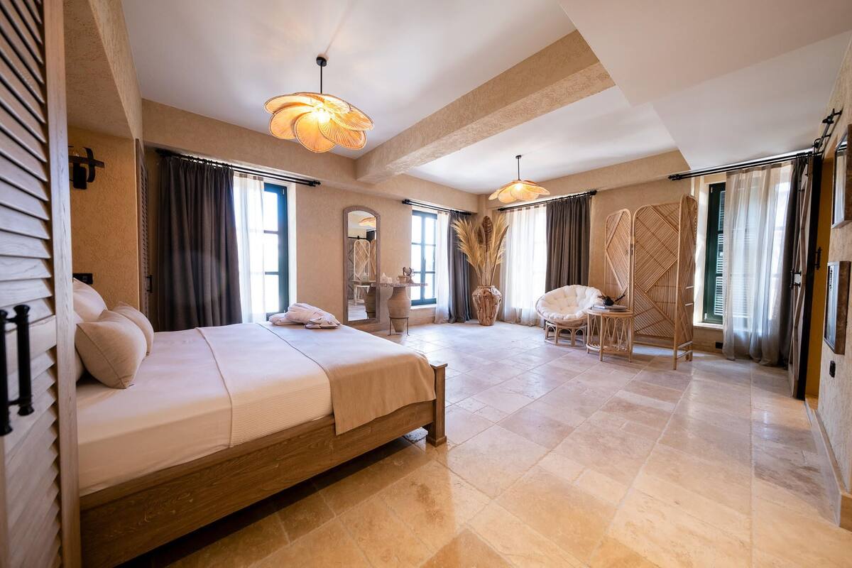 A spacious bedroom with a large bed, two pendant lights, a mirror, and a view of the outdoors.