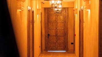 Un pasillo con una puerta de madera al final, flanqueado por otras puertas a ambos lados, y un candelabro colgando sobre él.