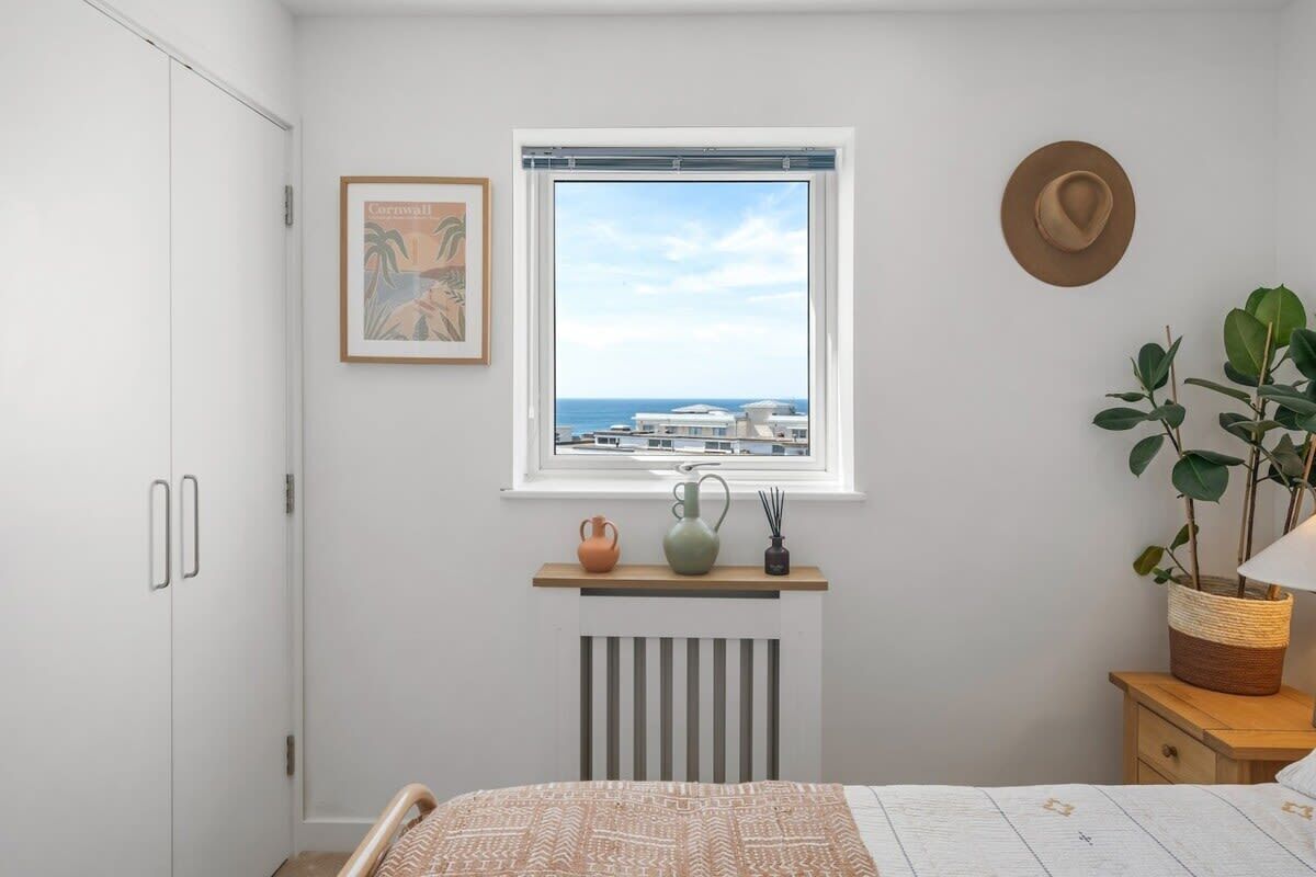 A bedroom with a bed, a window with a view, a radiator, a hat on the wall, and a framed picture.