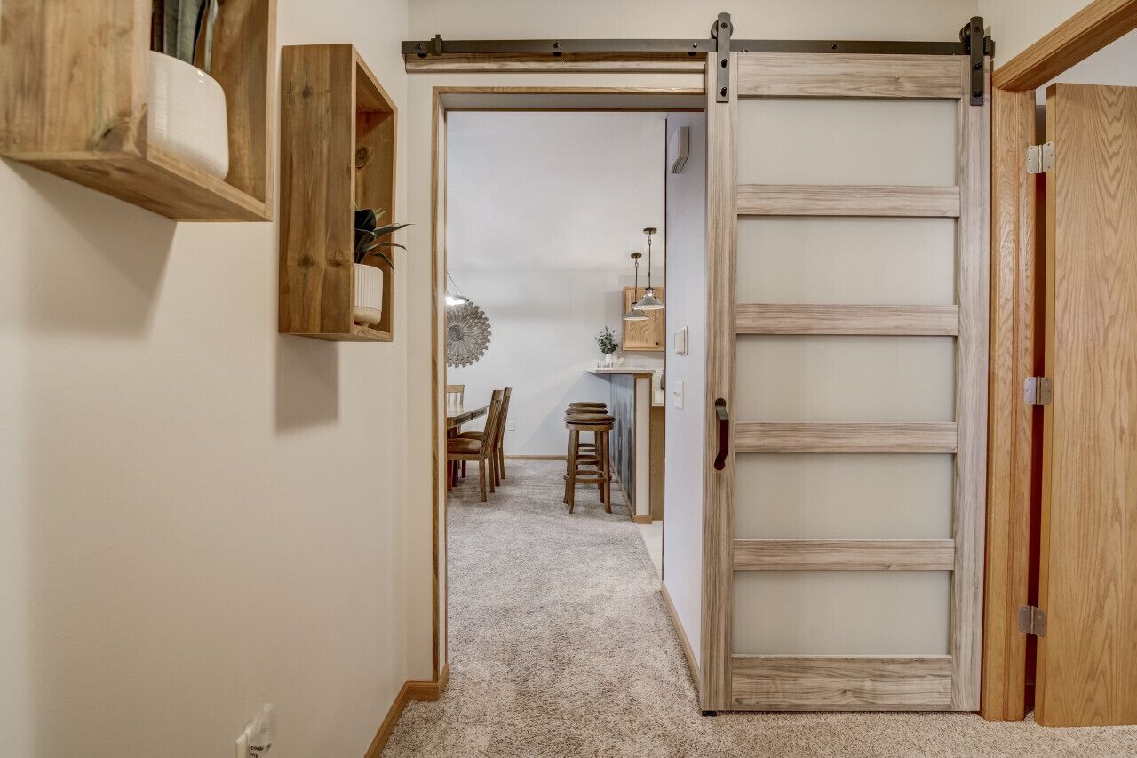 A hallway with a wooden door, a shelf with items, and a dining area in the background.