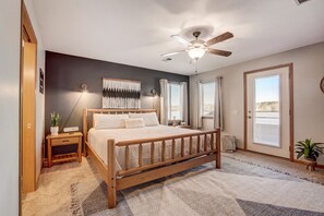 Une chambre à coucher avec un lit en bois, un ventilateur de plafond, une table de chevet, une lampe fixée au mur et une grande fenêtre avec des stores.