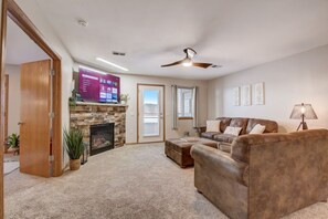 A living room with a stone fireplace, a leather sofa, a television mounted on the wall, and a ceiling fan. - Lakefront Haven - Families, Golfers, Girl's Trips (Wisconsin Dells)