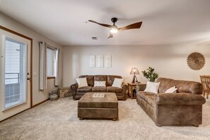 A living room with a brown leather sofa, a matching ottoman, a ceiling fan, and a large window with blinds. - Lakefront Haven - Families, Golfers, Girl's Trips (Wisconsin Dells)