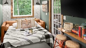 Een slaapkamer met een groot raam, een bed met een geruit sprei, een nachtkastje met boeken en een lamp, en een aan de muur bevestigde televisie.