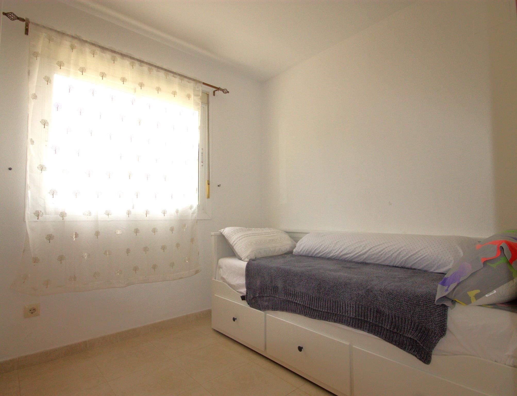Een slaapkamer met een bed, een raam met gordijn met boomprint en een witte muur.