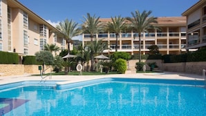 A swimming pool with a clear blue water, surrounded by palm trees and a multi-story building with balconies. - Golden - Javea, Costa Blanca (Javea)
