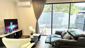 Una sala de estar moderna con un sofá de cuero, un sillón blanco, un televisor sobre un mueble bajo y un ventanal con vistas a un jardín.