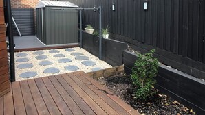 Una terraza de madera con piedras que forman un camino hacia un cobertizo, rodeado por una cerca negra y un muro de ladrillo.